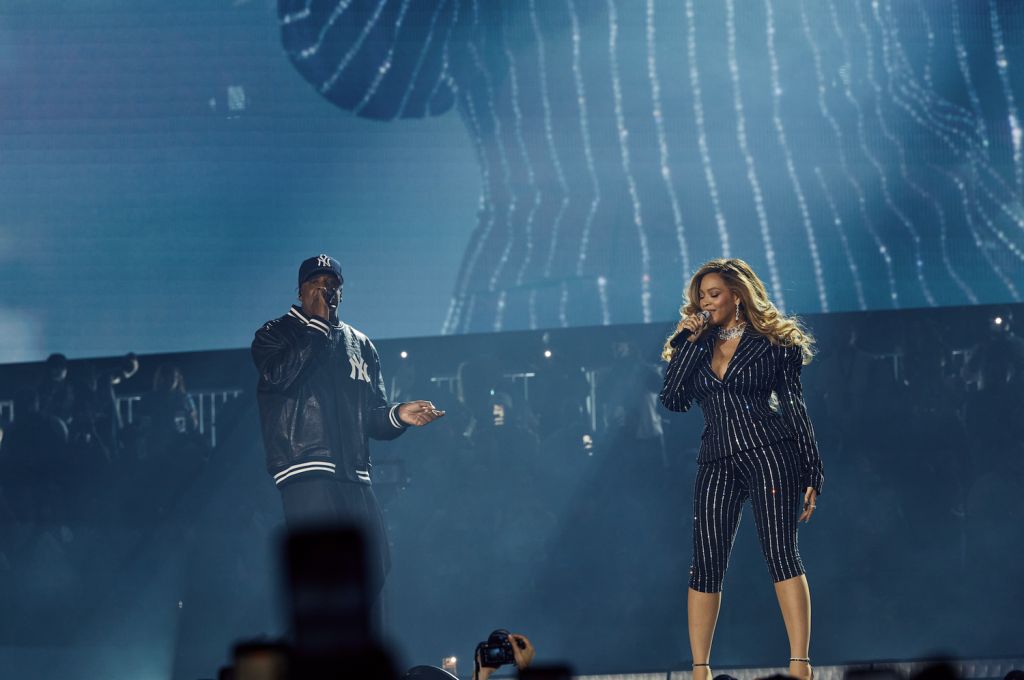 Two performers on stage, a man in a Yankees baseball cap and a woman in a black and white striped jumpsuit, singing and dancing under bright lights.