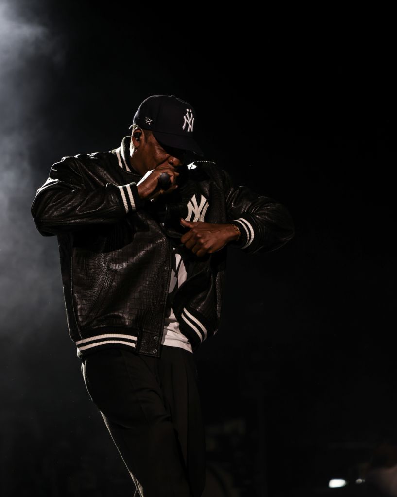 A person wearing a New York Yankees baseball cap and jacket, standing in a dark, smoky setting and holding a microphone.