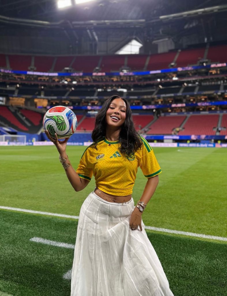 A smiling woman in a yellow soccer jersey holding a multicolored soccer ball on a soccer field in a large stadium.
