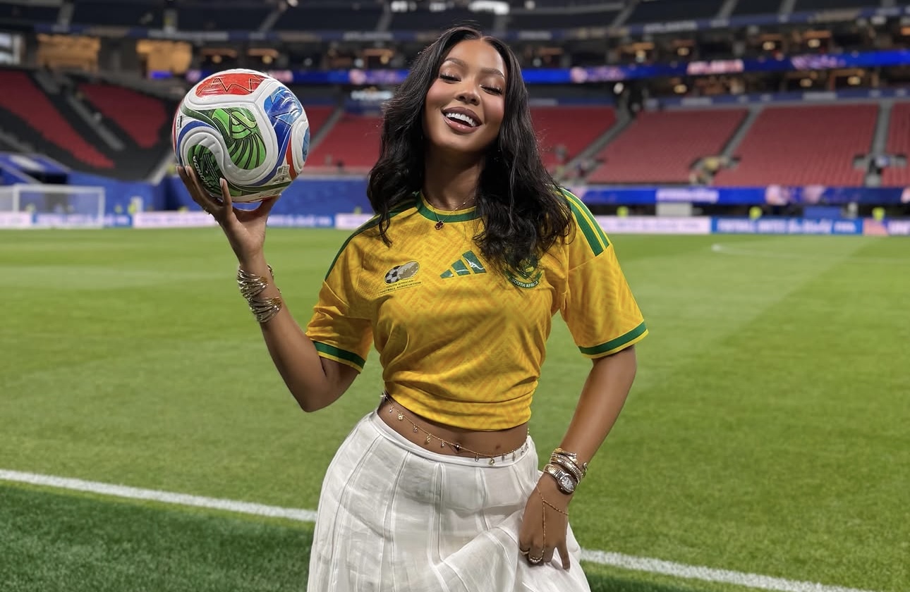 A smiling woman in a yellow soccer jersey holding a soccer ball on a soccer field.