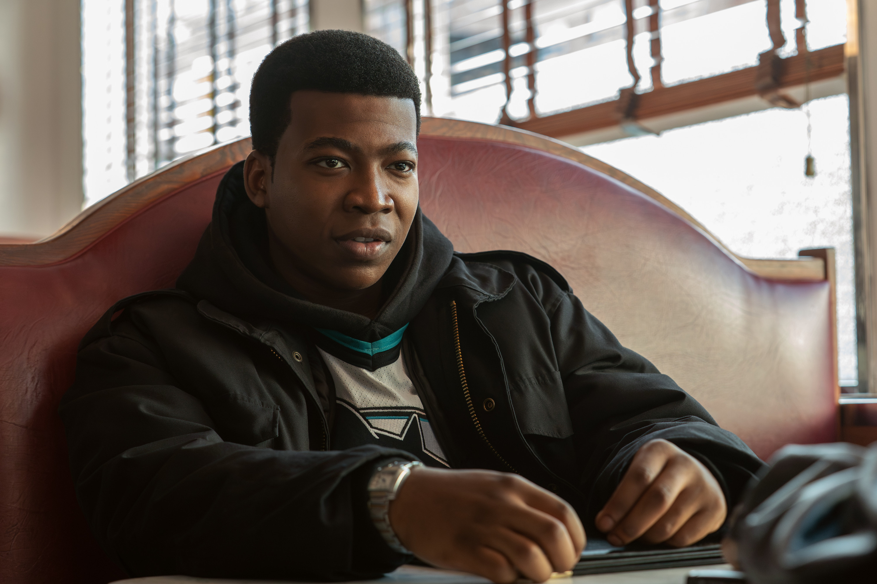 A young Black man sitting in a chair, wearing a black jacket and looking directly at the camera with a serious expression.