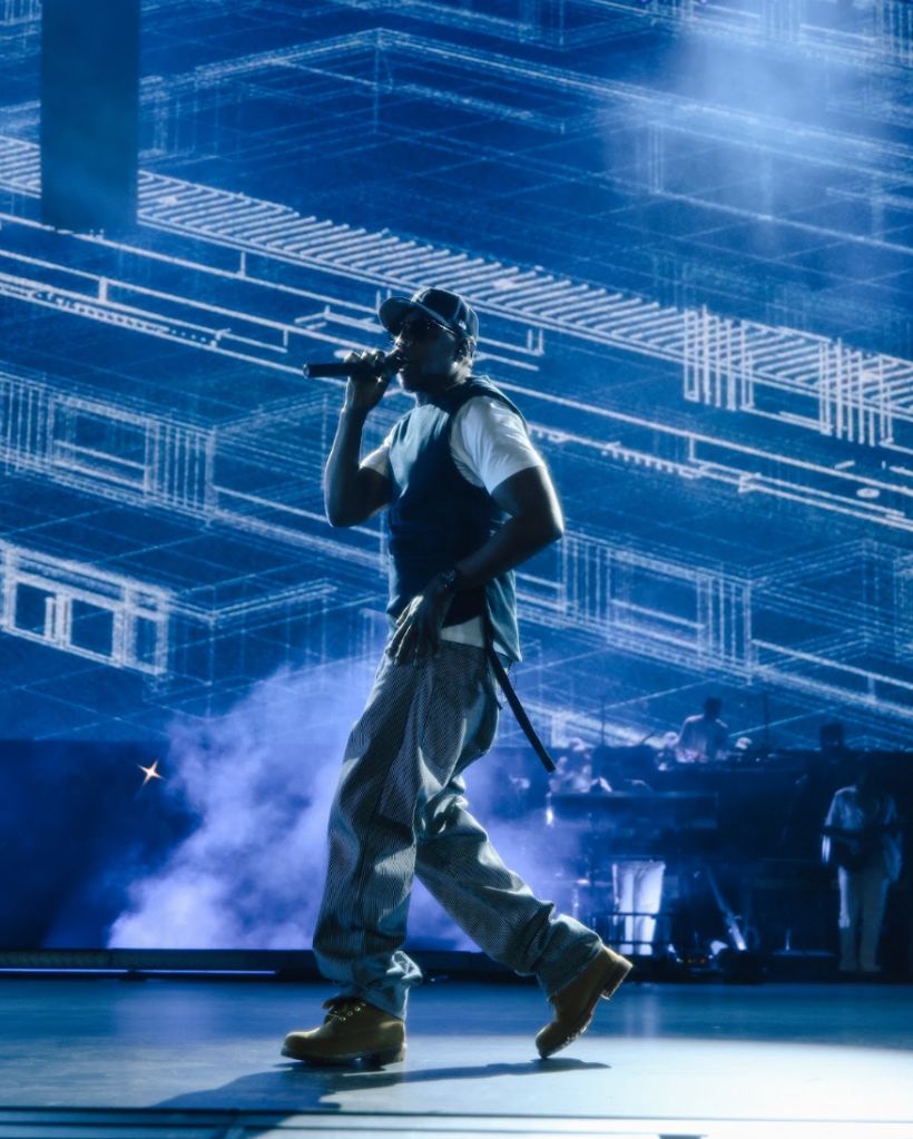 A performer on stage in a dark, futuristic setting, wearing a white shirt and dark pants, holding a microphone and surrounded by smoke and blue lighting.