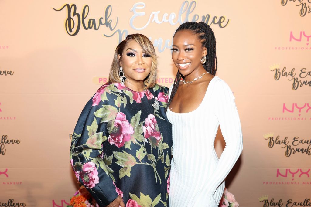 Two women posing in front of a backdrop with "Black Excellence" text. One woman wears a floral print dress, the other a white dress with long sleeves.
