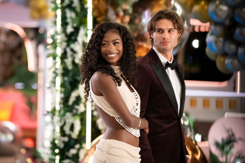 Two people, a smiling woman with curly hair and a man in a tuxedo, standing together in front of a festive holiday backdrop with lights and greenery.