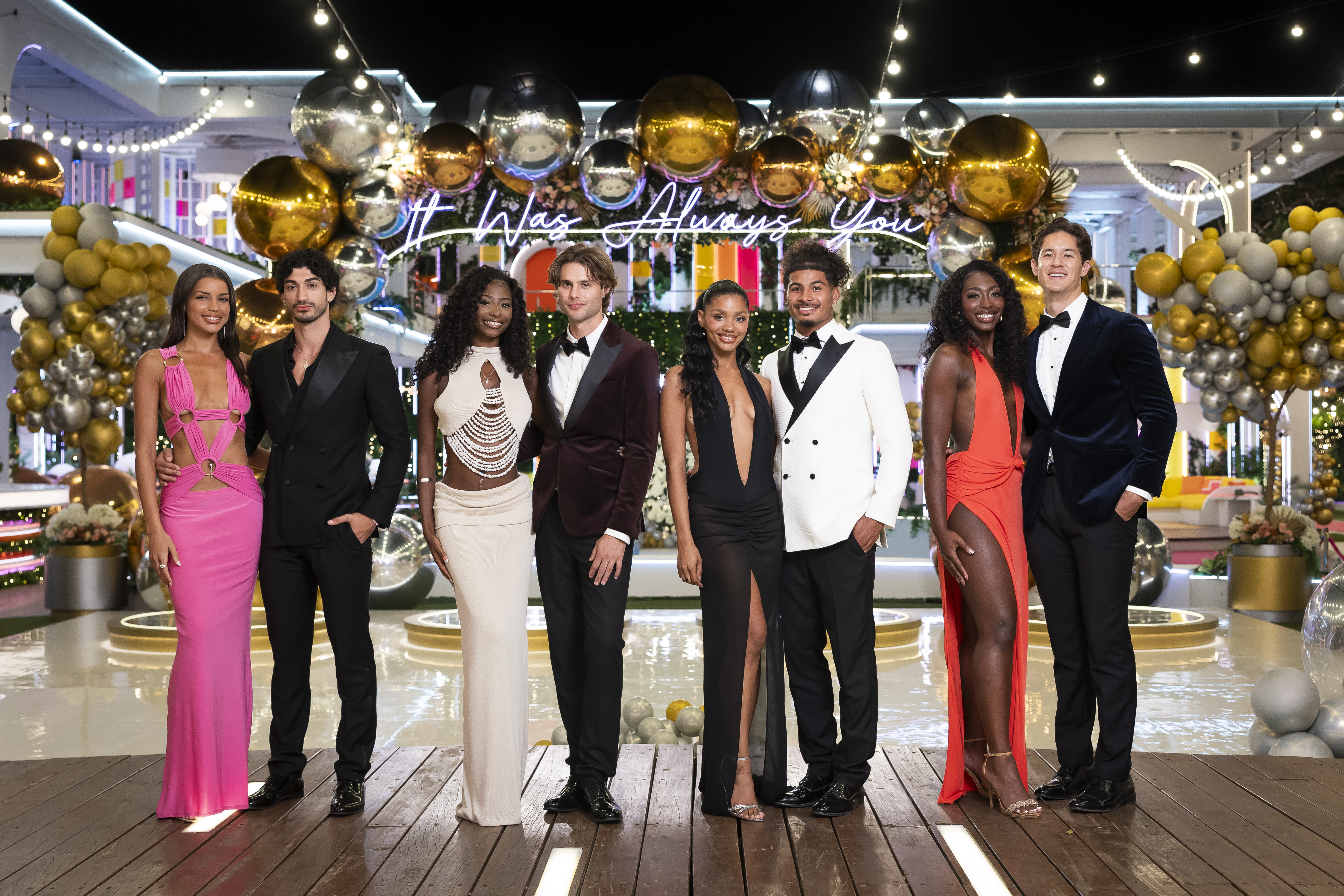 A group of well-dressed people posing in front of a festive backdrop with gold and silver balloons and the text "It was always you".