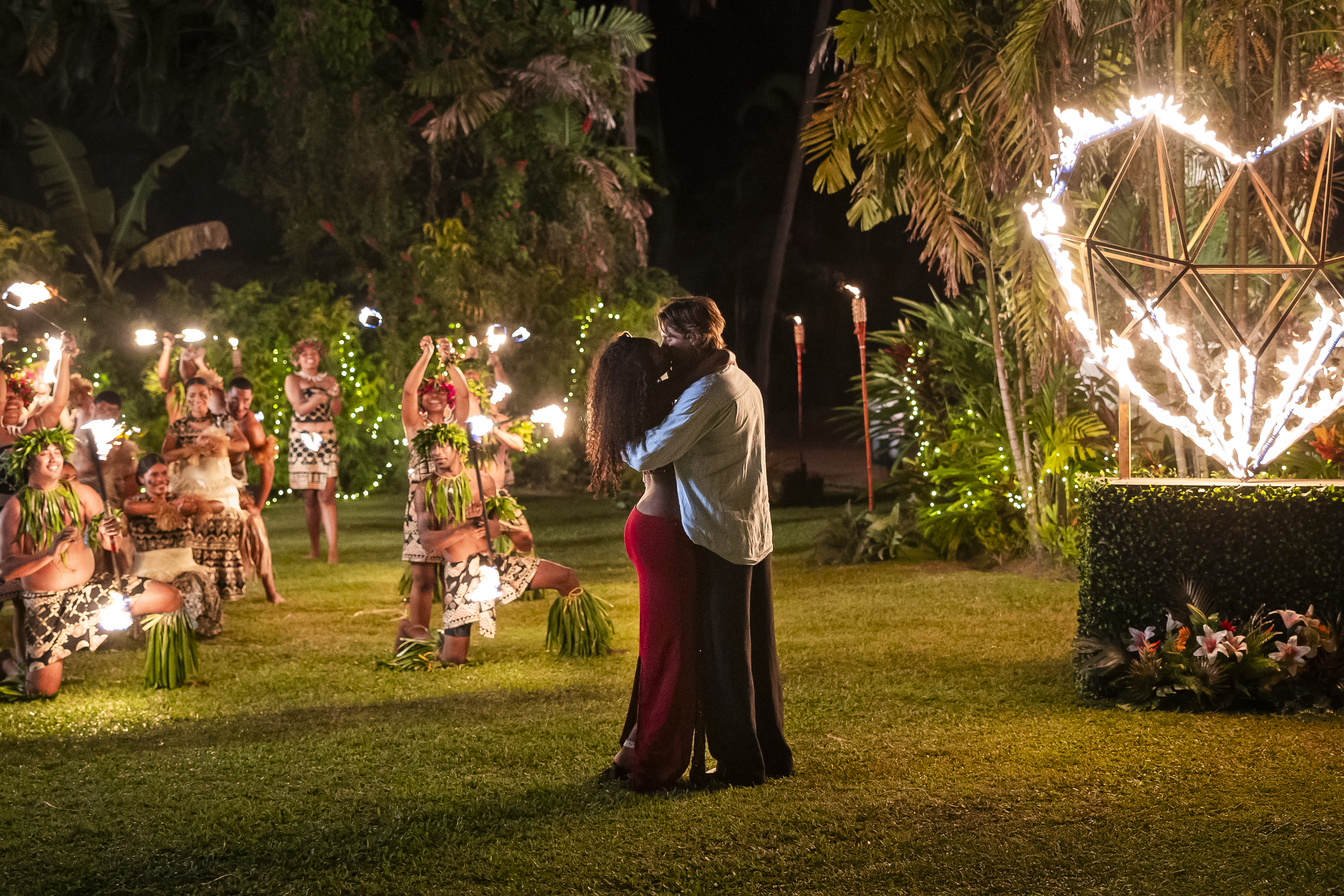 A nighttime tropical garden scene with a couple embracing and dancers in traditional costumes performing amidst twinkling lights and palm trees.