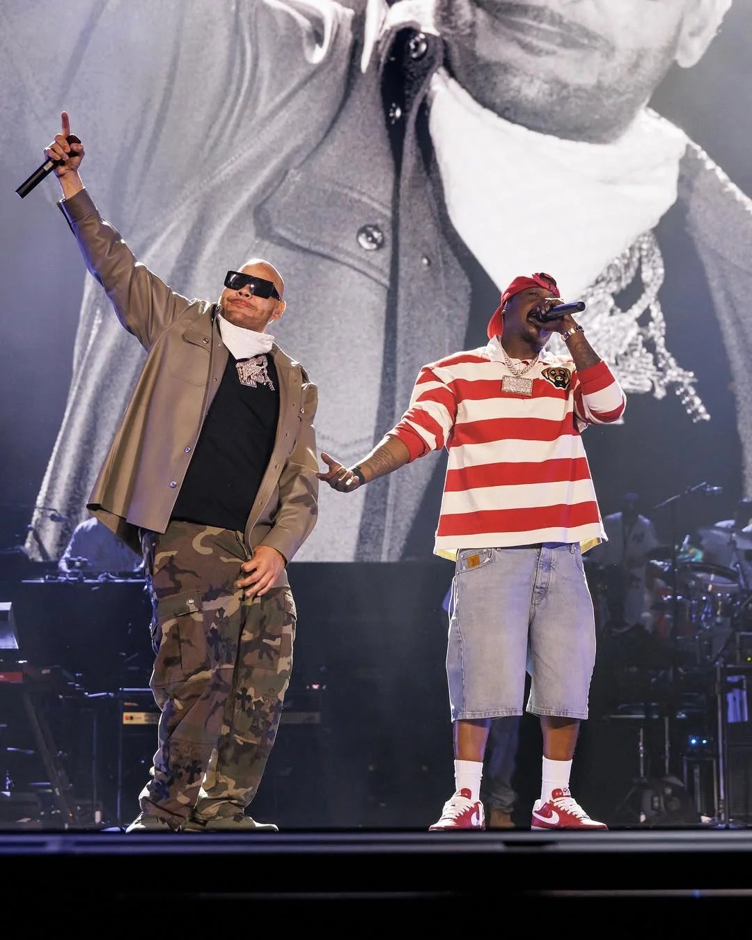 Two performers on stage, one wearing camouflage pants and a black jacket, the other in a red and white striped shirt, both holding microphones and performing.