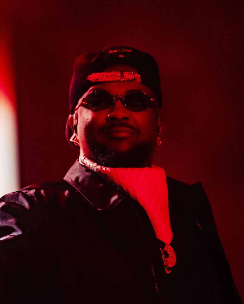 A person wearing a black cap, sunglasses, and a red shirt, standing in a dimly lit setting with a red light.