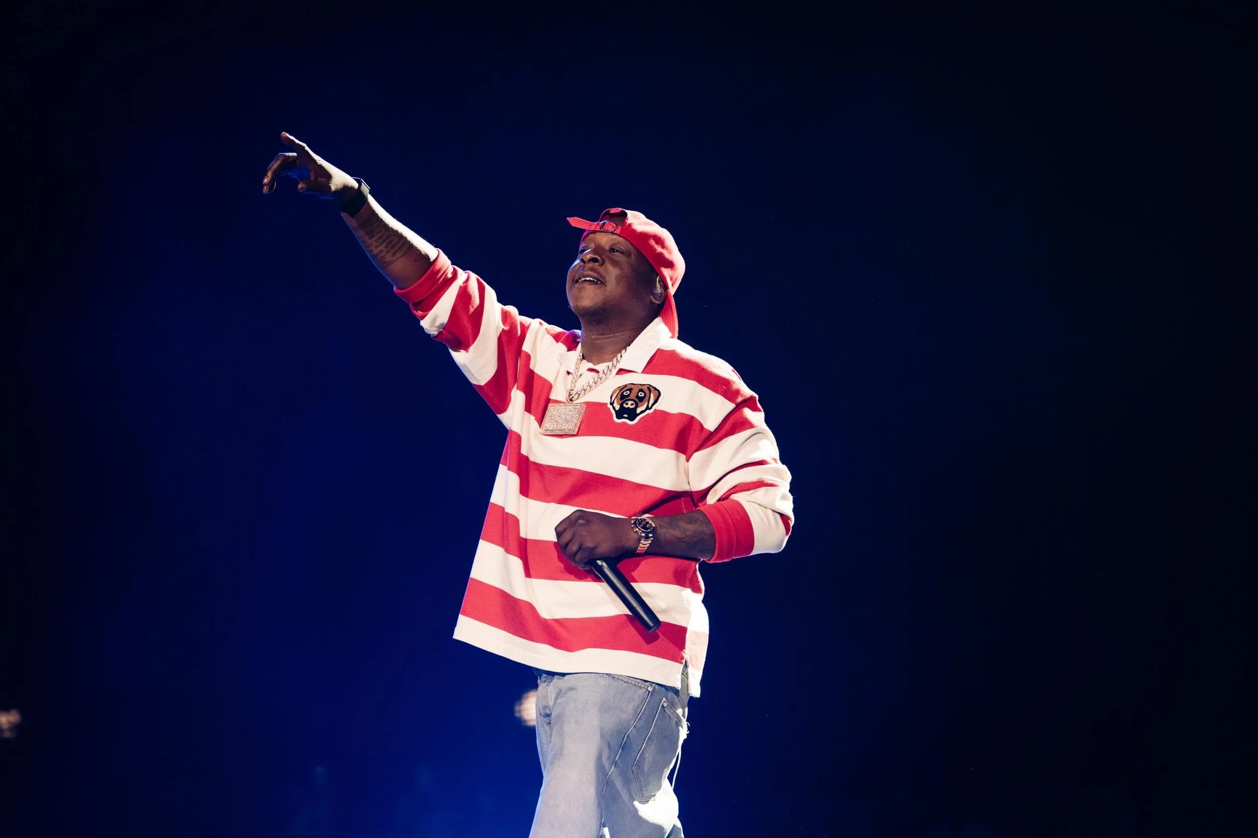 A performer in a red and white striped jersey and red cap gesturing on a dark stage.