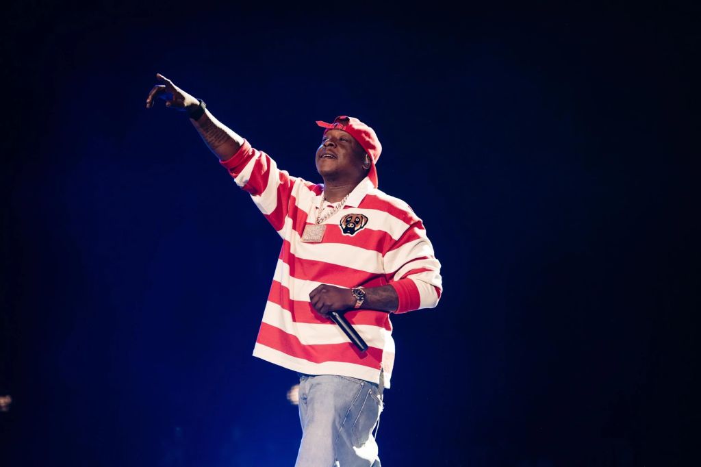 A performer in a red and white striped jersey and red cap gesturing on a dark stage.