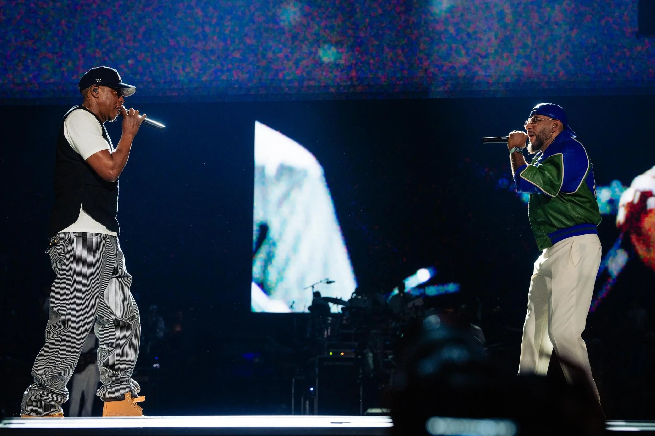 Two musicians performing on stage, one wearing a black cap and the other a green jacket, singing into microphones against a colorful backdrop.