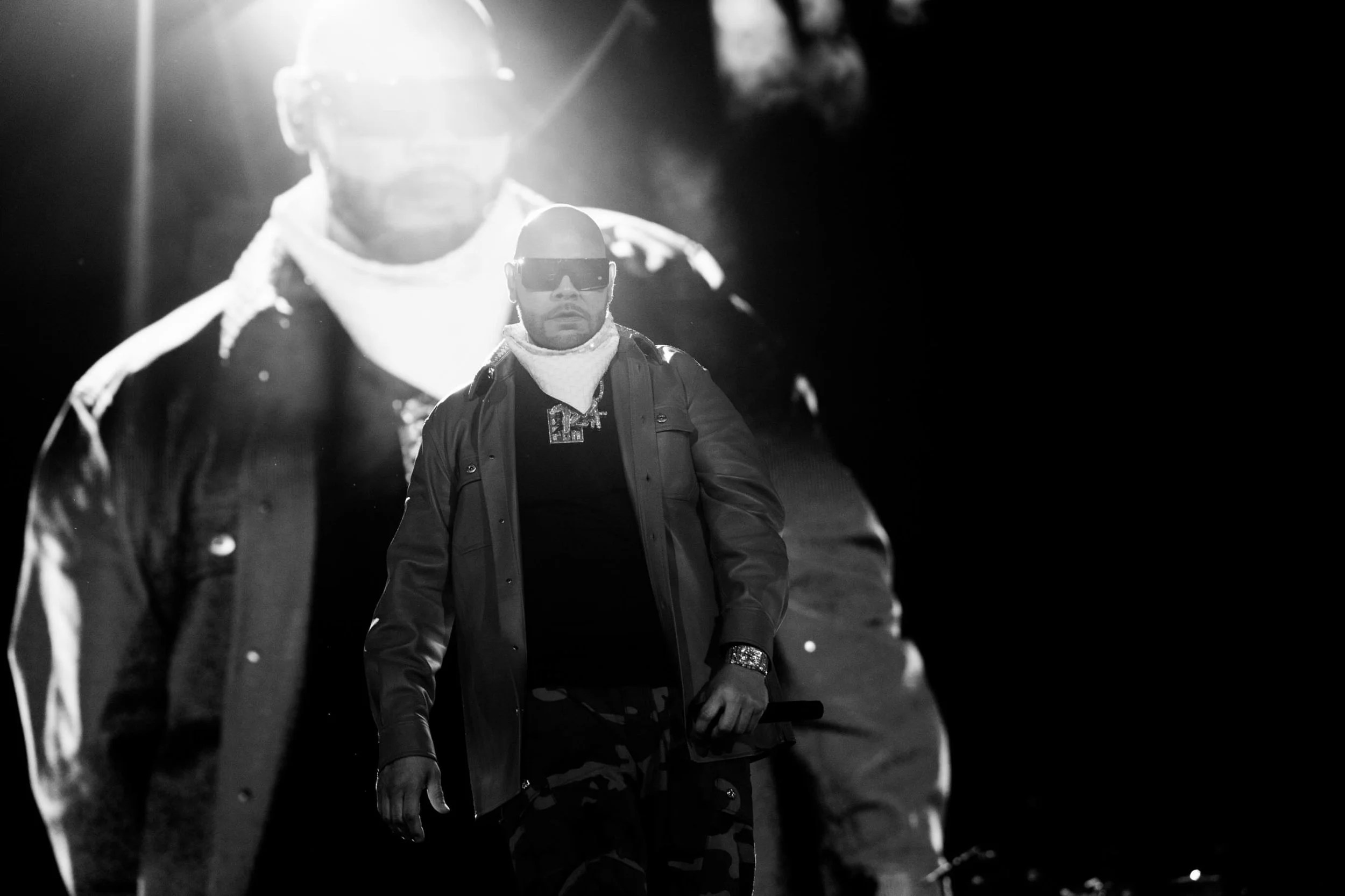 A person wearing a black jacket and sunglasses standing in a dimly lit environment with a bright light source behind them.