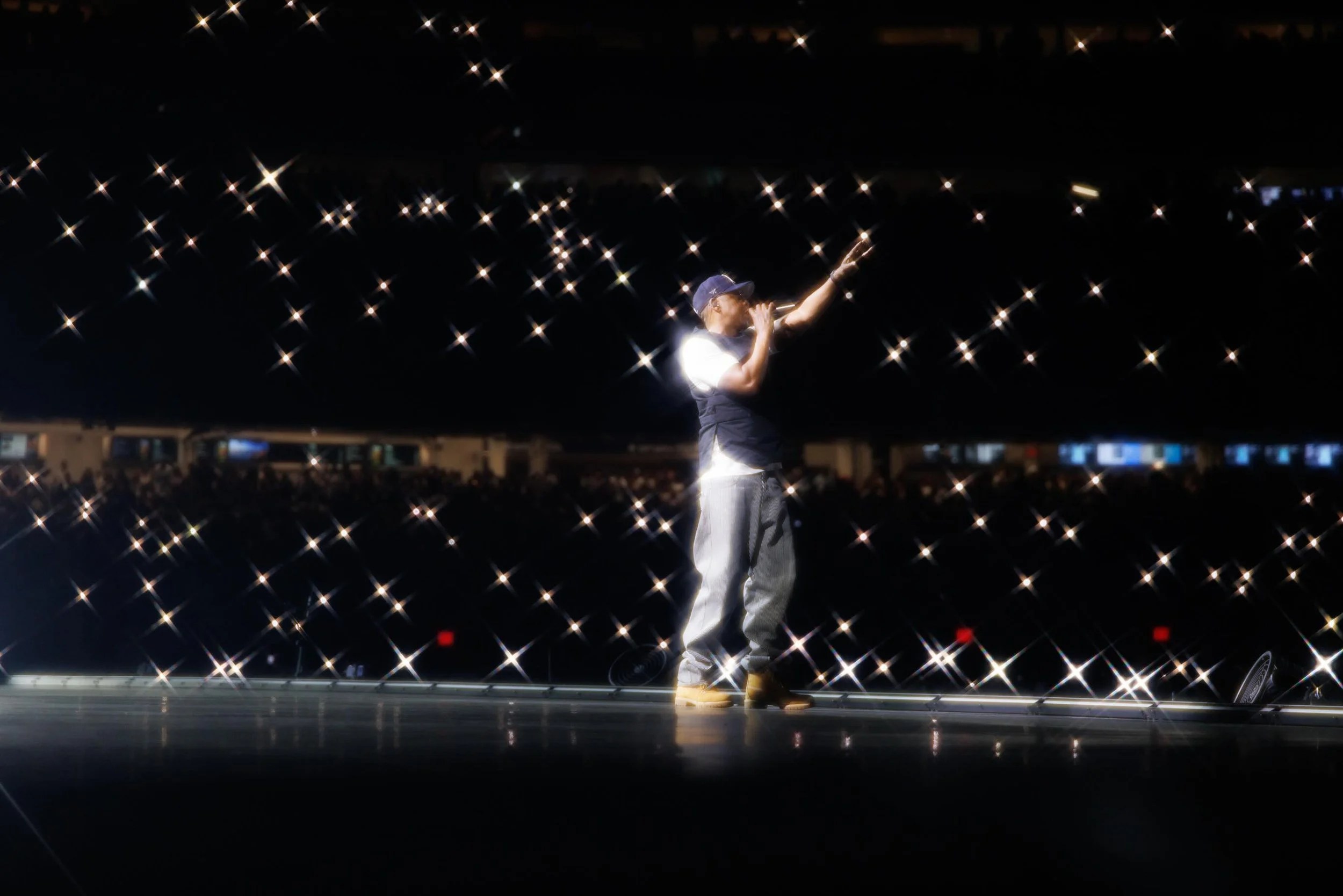 A performer on stage at night, arms raised, surrounded by twinkling lights in the dark sky.