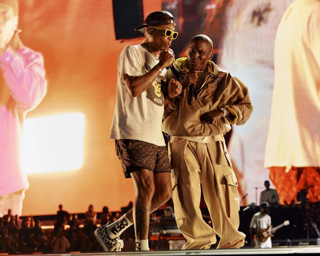 Two people on stage, one wearing a white t-shirt with "Made" printed on it and sunglasses, the other wearing a beige jacket, performing together.