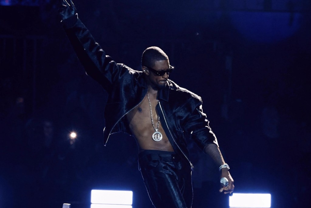 A shirtless man wearing sunglasses and a necklace performing on stage in a dark, dramatic lighting.