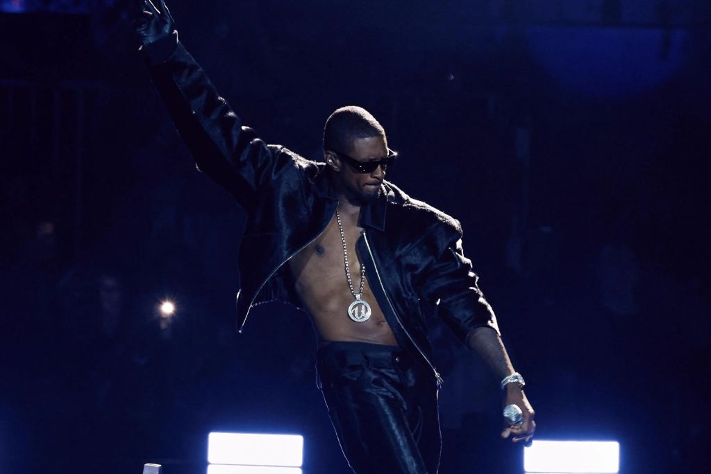A shirtless man wearing sunglasses and a necklace performing on stage in a dark, dramatic lighting.