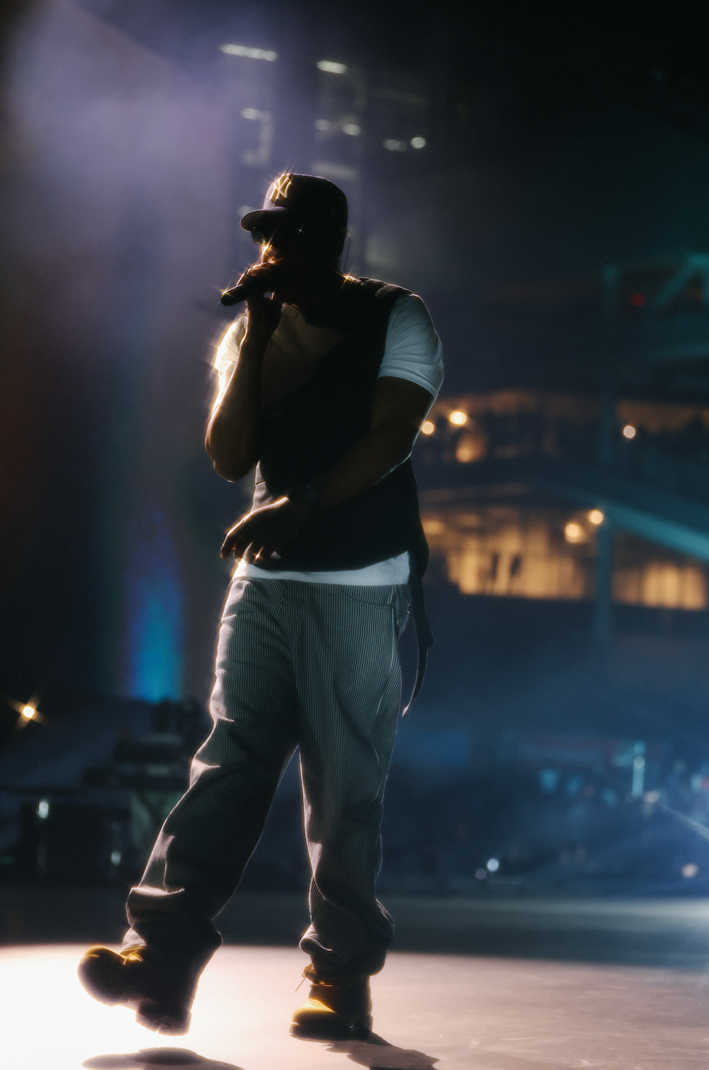 A person wearing a black cap and shirt performs on a dimly lit stage, with lights and shadows creating a dramatic effect.
