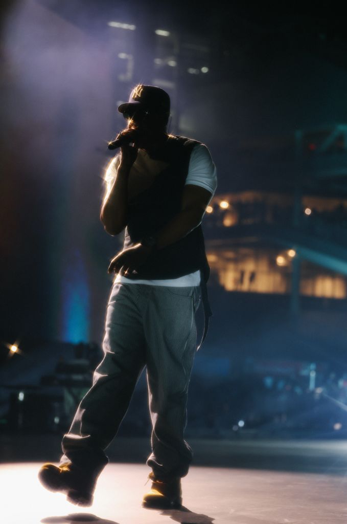 A person wearing a black cap and shirt performs on a dimly lit stage, with lights and shadows creating a dramatic effect.