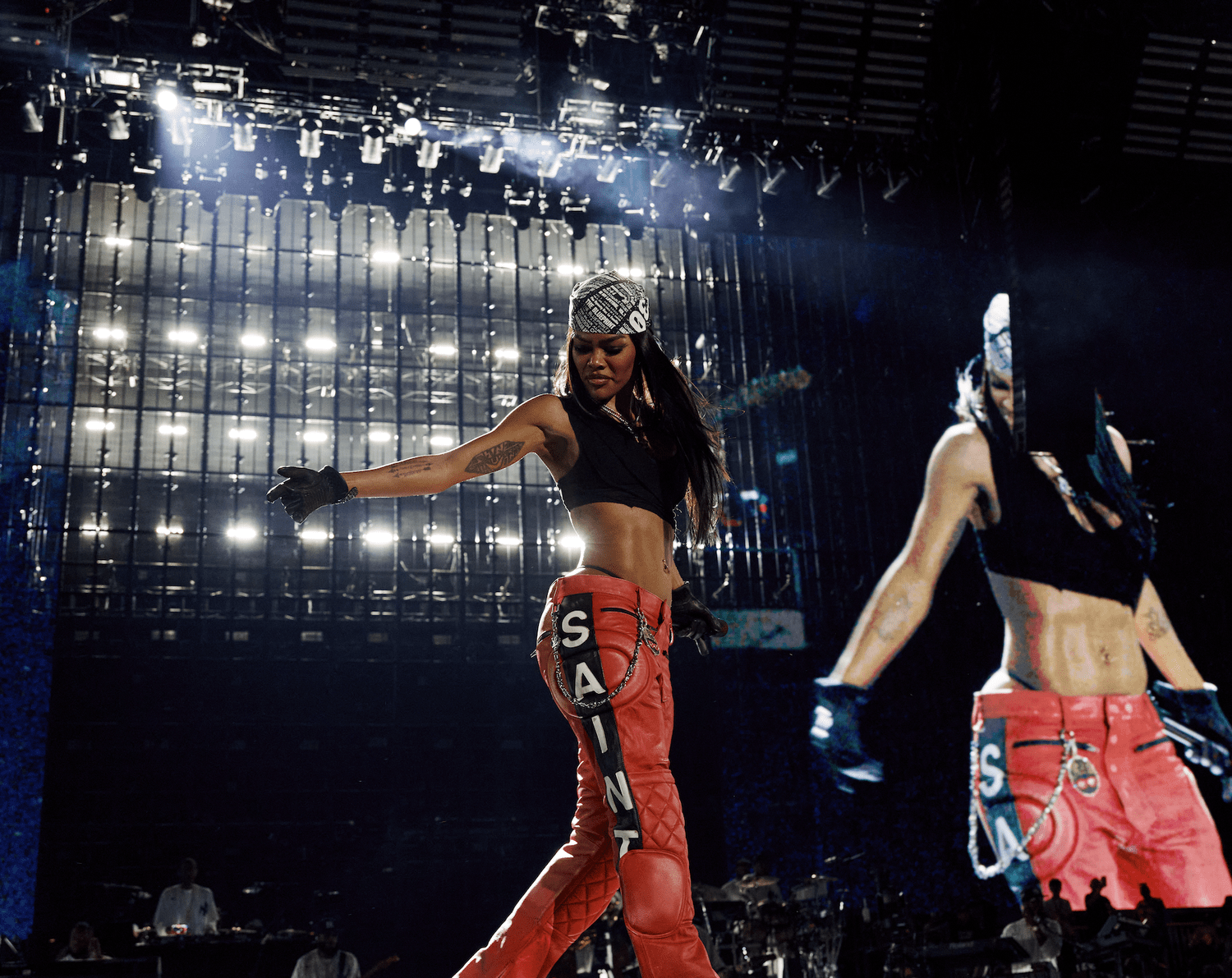 A performer wearing a black crop top and red quilted pants dances on a stage with bright lights and a backdrop of the word "SANA".