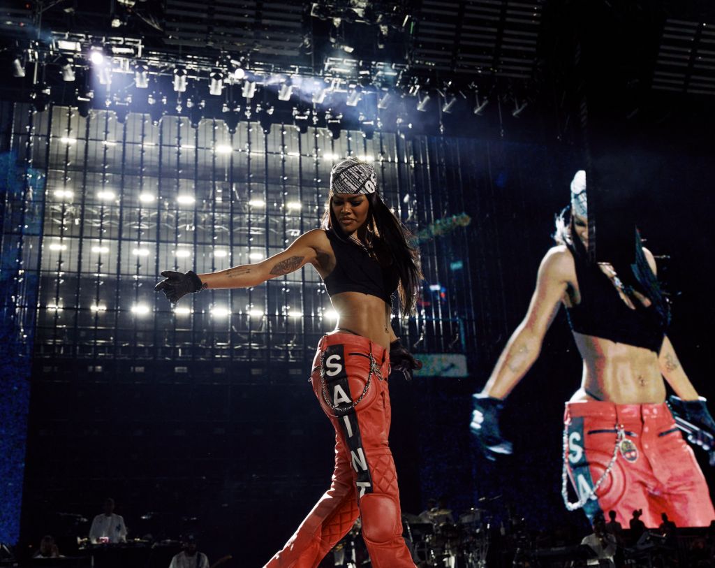 A performer wearing a black crop top and red quilted pants dances on a stage with bright lights and a backdrop of the word "SANA".