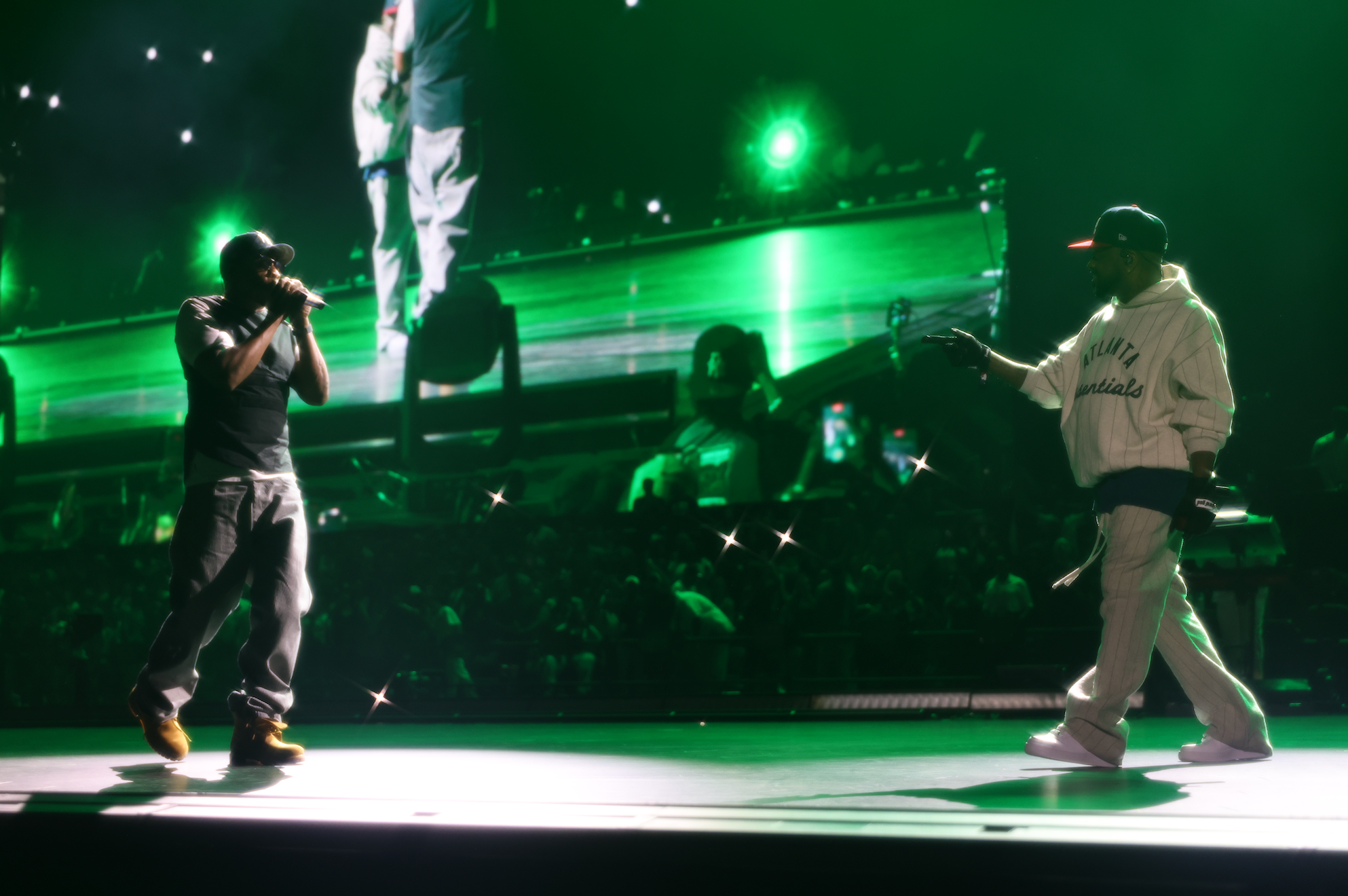 Two performers on stage in a dimly lit concert venue, one singing into a microphone and the other dancing, with green stage lighting illuminating the scene.