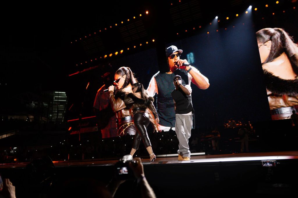 Two performers on stage, one wearing a leather jacket and sunglasses, the other in a red outfit, singing and dancing before a backdrop of lights.