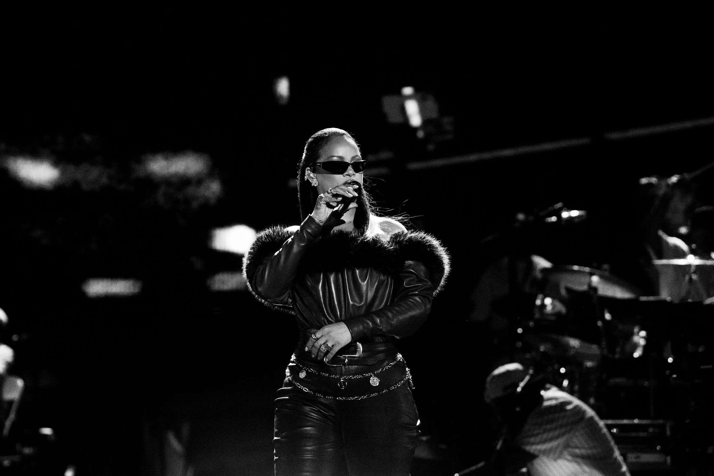 A person wearing sunglasses and a fur-trimmed jacket performing on stage in front of a drum set.