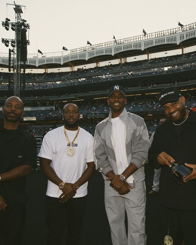 Four men standing together in a baseball stadium, smiling and wearing casual clothing. The New York Yankees logo is visible on the stadium.