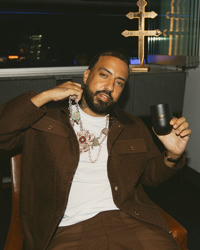 A man with a beard wearing a brown jacket and holding a mug, with a cross-shaped object visible in the background.