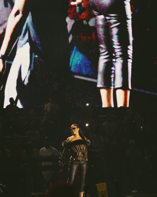 A performer wearing sunglasses and a black outfit with a blue and white pattern stands on a dark stage with colorful lights and graphics projected behind them.