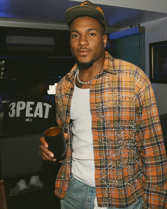 A man wearing a plaid shirt, cap, and necklace standing in front of a "3PEAT" backdrop.