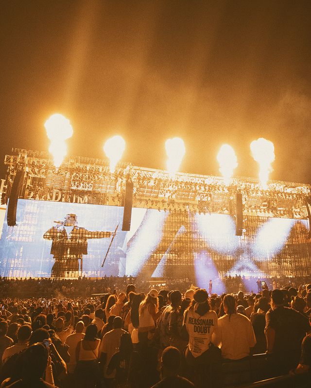 A large crowd at a music festival or concert, with bright lights and pyrotechnics illuminating the stage and audience. The text "REASONABLE DOUBT" is visible on a person's shirt in the foreground.