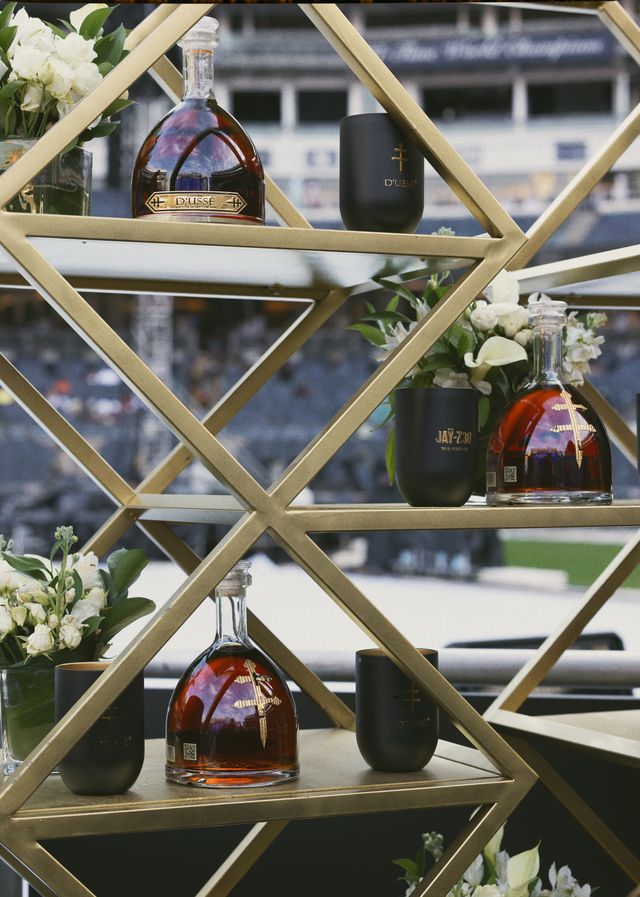 Shelves displaying bottles of Dusse and Jay-Z cognac, surrounded by white flowers in a decorative metal frame.