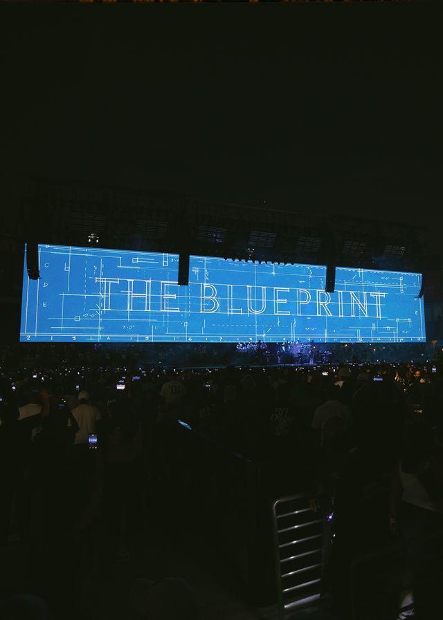 A large projection screen displaying the text "THE BLUEPRINT" against a dark background, with a crowd visible in the foreground.