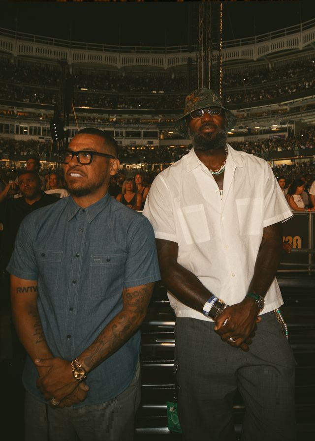 Two men, one wearing glasses and a denim shirt and the other wearing a white shirt and sunglasses, standing in a crowded arena.