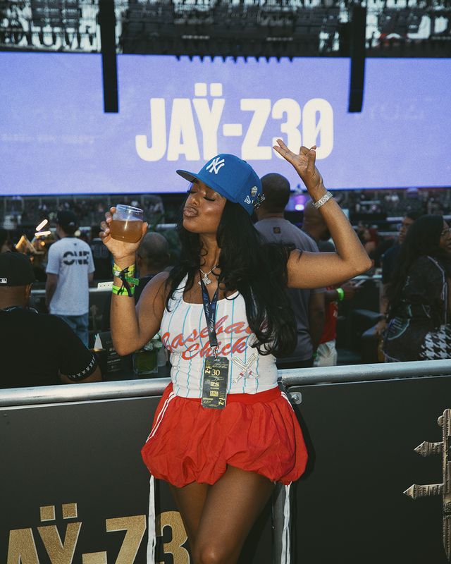 A person wearing a Yankees cap and a red skirt dancing and holding a drink in front of a large "JAY-Z 730" screen.