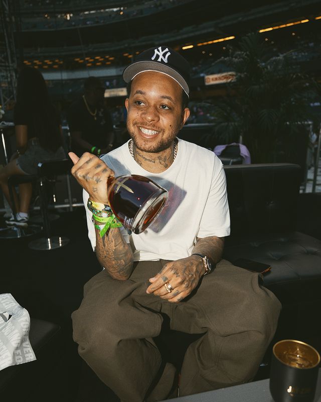 A smiling man wearing a Yankees cap and holding a drink, seated in a dimly lit venue.