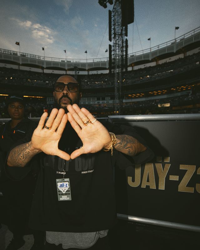 A man with tattoos and sunglasses making a hand gesture in a large sports stadium, with the text "JAY-Z" visible on his shirt.