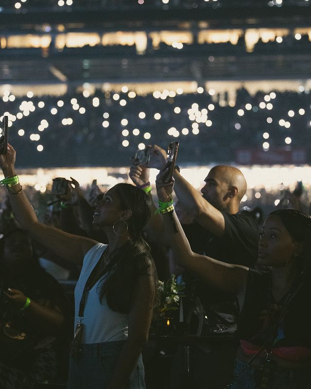 Crowd of people with raised hands and phones at a nighttime event, with bright lights and bokeh in the background.