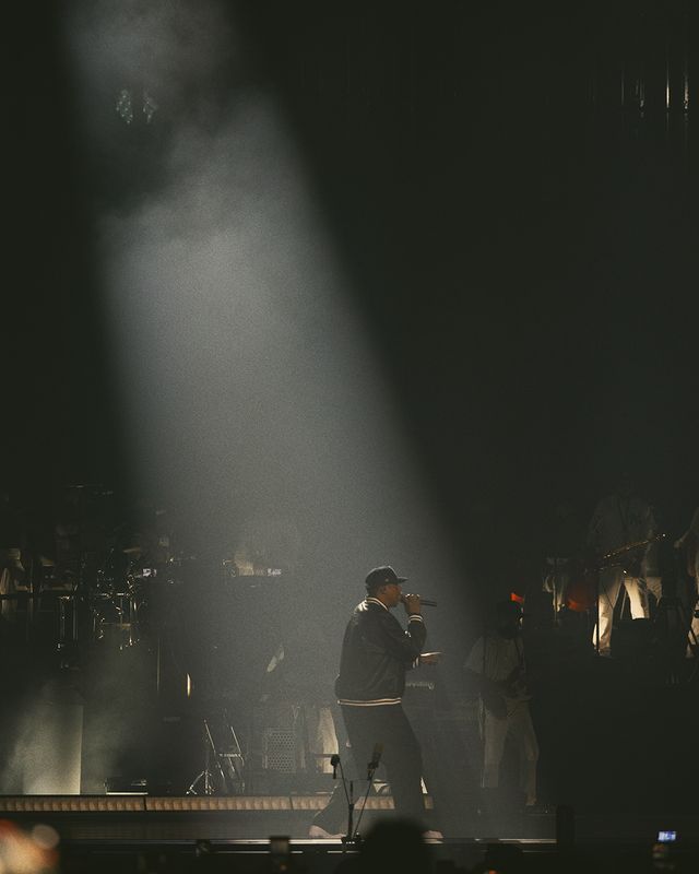 A dimly lit stage with a performer silhouetted against a bright spotlight, surrounded by a crowd of people.