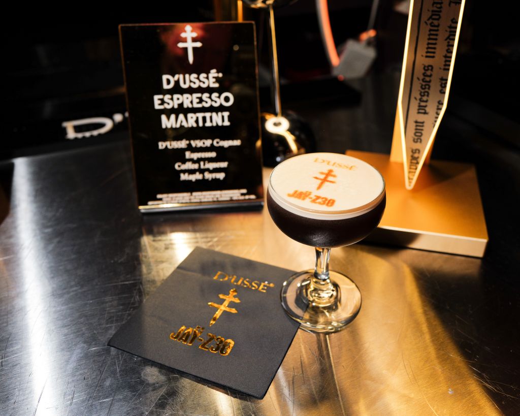 A glass of dark espresso martini on a table, with a bottle and menu for "D'usse Espresso Martini" visible.