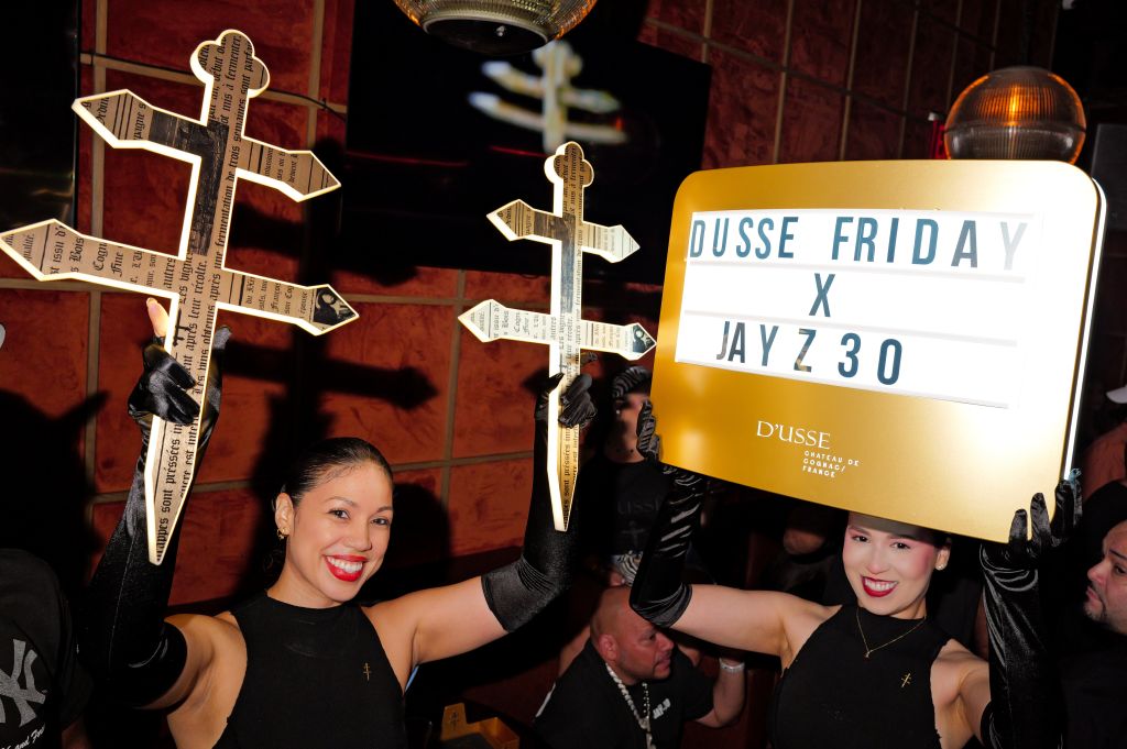 Two women holding large illuminated crosses, with a sign advertising "Dusse Friday x Jay Z 30" event.
