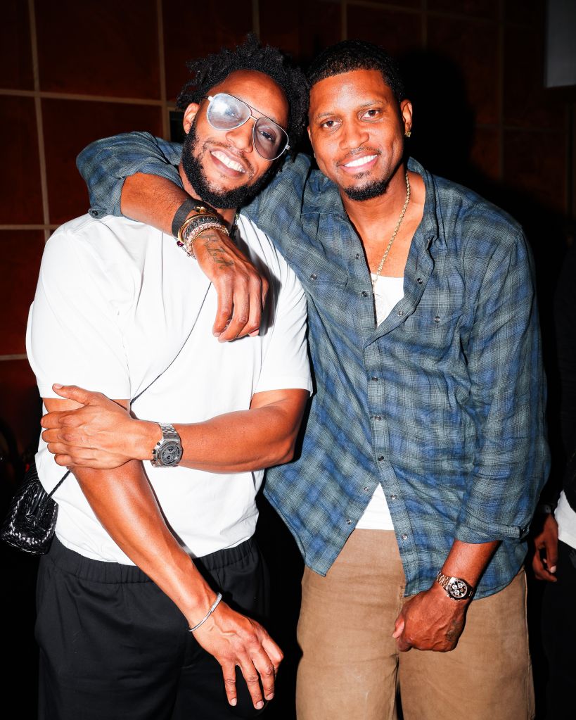 Two smiling men wearing casual clothing and accessories, embracing each other.