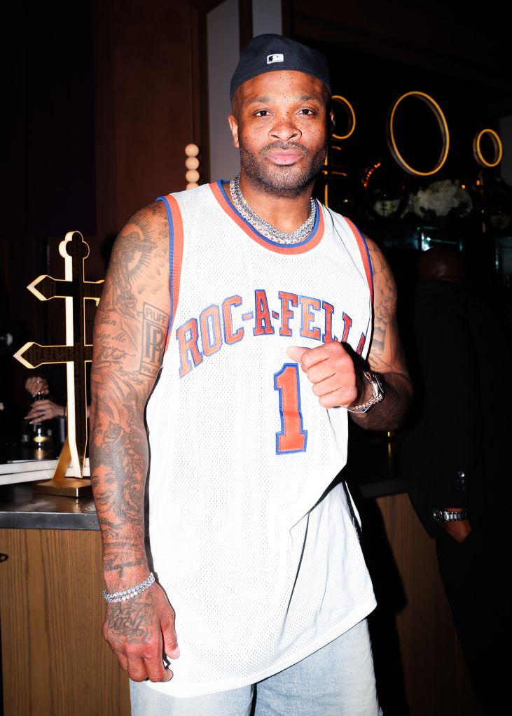 A man wearing a white basketball jersey with "ROC-A-FELLA 1" printed on it, standing in a room with decorative lighting.