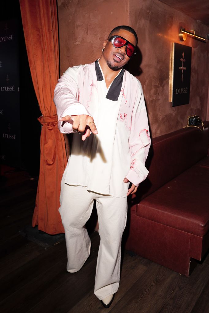 A person wearing a white suit with pink splatter patterns, red sunglasses, and a black tie, standing in a room with orange curtains and a Dusse logo visible.