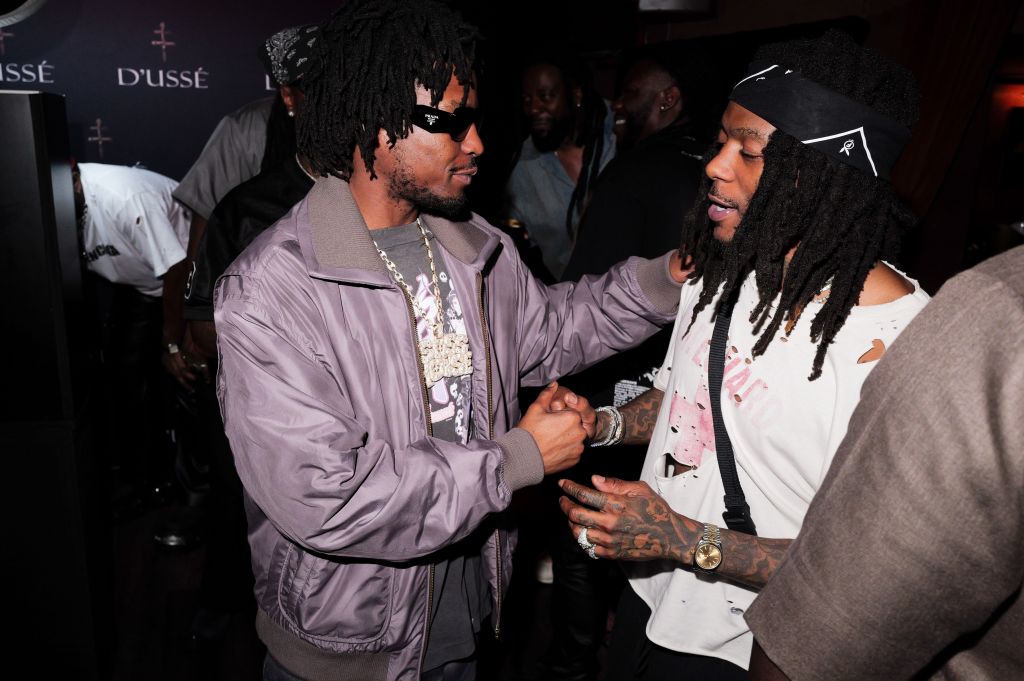 Two men in casual clothing, one wearing a gray jacket, the other with dreadlocks, shaking hands and embracing.