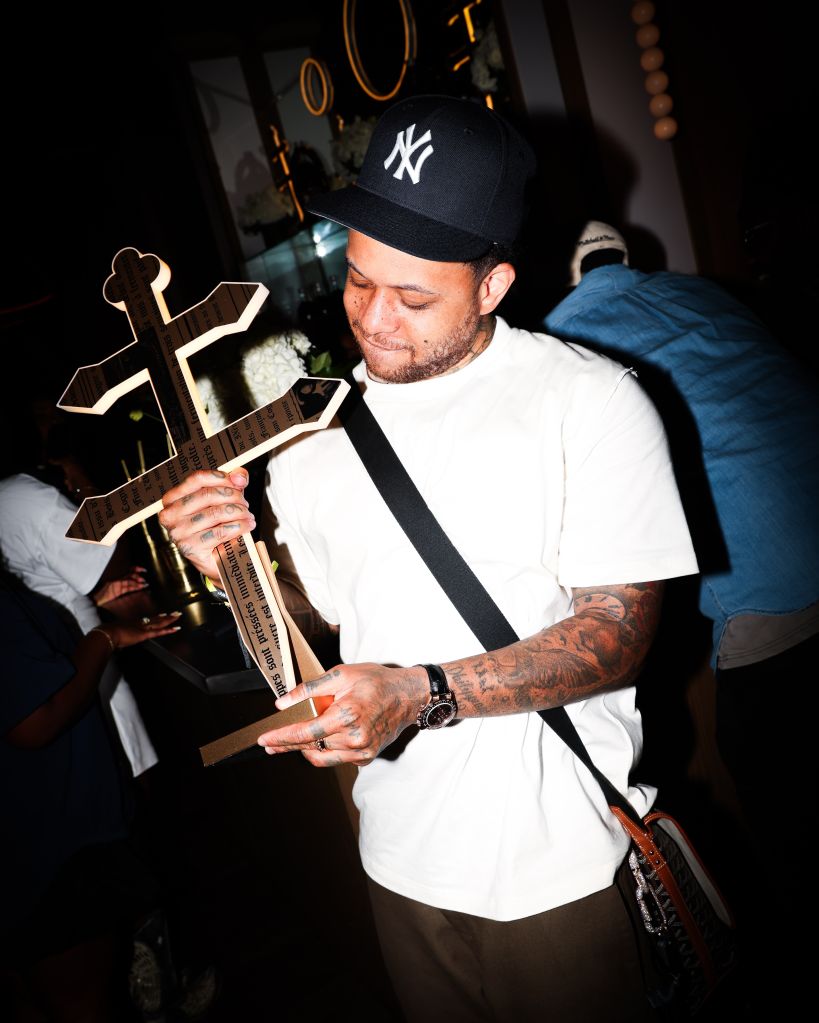 A man wearing a New York Yankees cap and holding a large cross-shaped object, with tattoos visible on his arms.