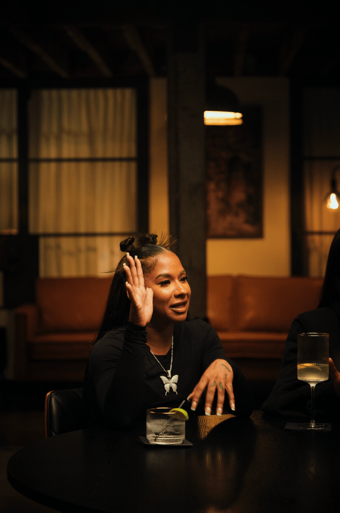 A person sitting at a table in a dimly lit room, smiling and gesturing with their hand.