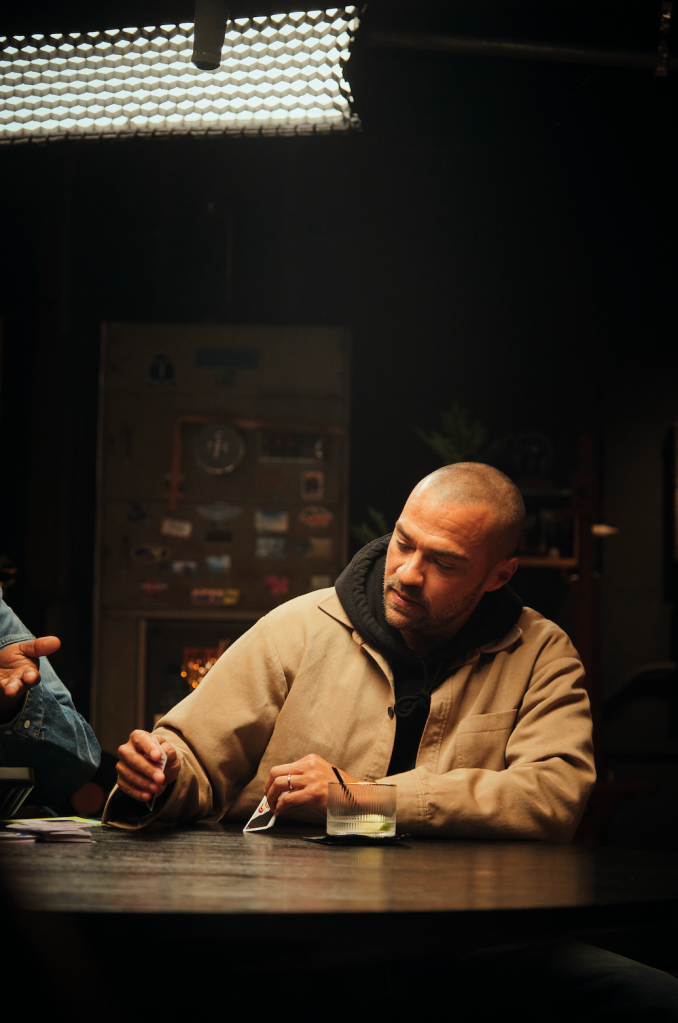 A man sitting at a table in a dimly lit room, looking down at something in his hands.