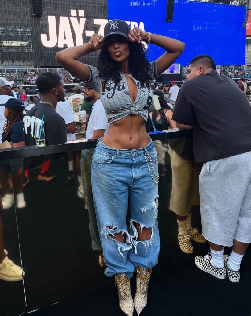 A woman wearing a Yankees cap and crop top poses in front of a large "JAY-Z" sign at what appears to be a music or entertainment event.